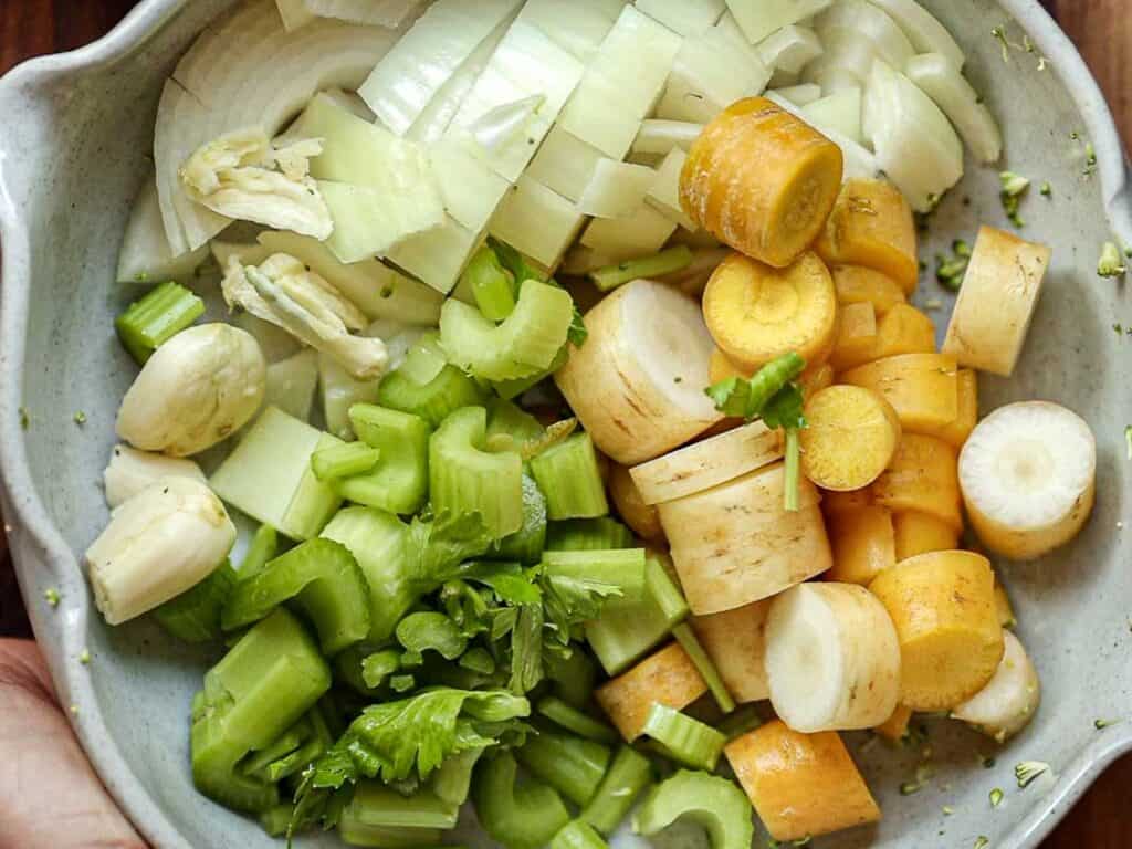 A bowl filled with chopped vegetables, including onions, yellow carrots, garlic cloves, and celery, arranged in sections. The ingredients are fresh and colorful, highlighting a mix of greens, yellows, and whites.